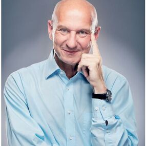 Ein älterer Mann in hellblauem Hemd lächelt selbstbewusst vor grauem Hintergrund.