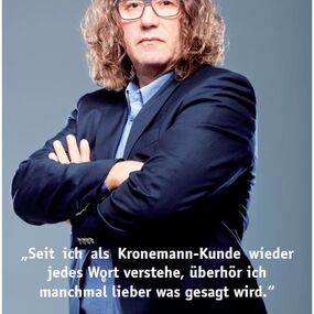Mann mit langen Haaren und Brille steht im Anzug vor grauem Hintergrund mit Zitat.